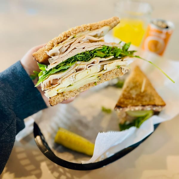 A hand holds a turkey and brie sandwich with arugula, next to a pickle spear and drink.