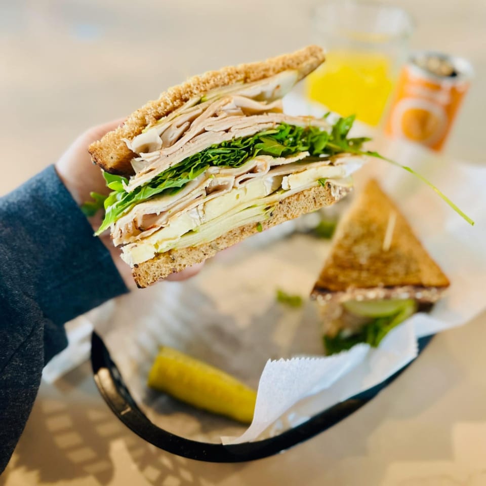 A hand holds a turkey and brie sandwich with arugula, next to a pickle spear and drink.