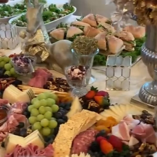A deli spread featuring sandwiches, charcuterie, cheeses, grapes, and other snacks on a white tablecloth.