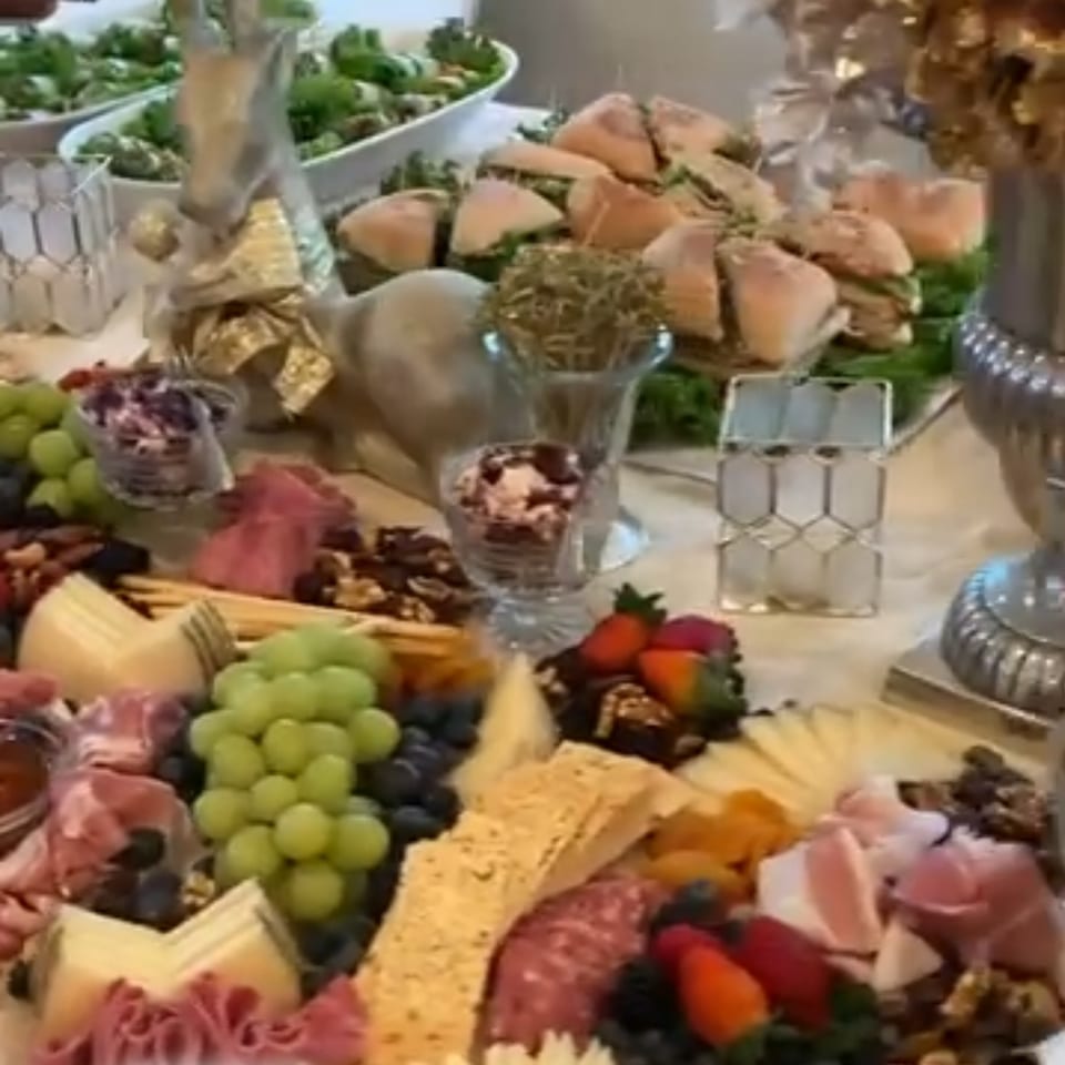 A deli spread featuring sandwiches, charcuterie, cheeses, grapes, and other snacks on a white tablecloth.