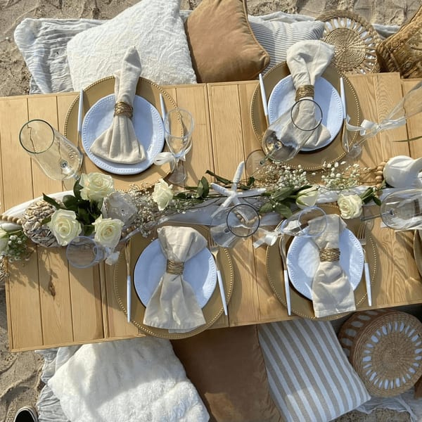 A beach picnic setup with a low wooden table, place settings, white roses, and pillows on the sand.