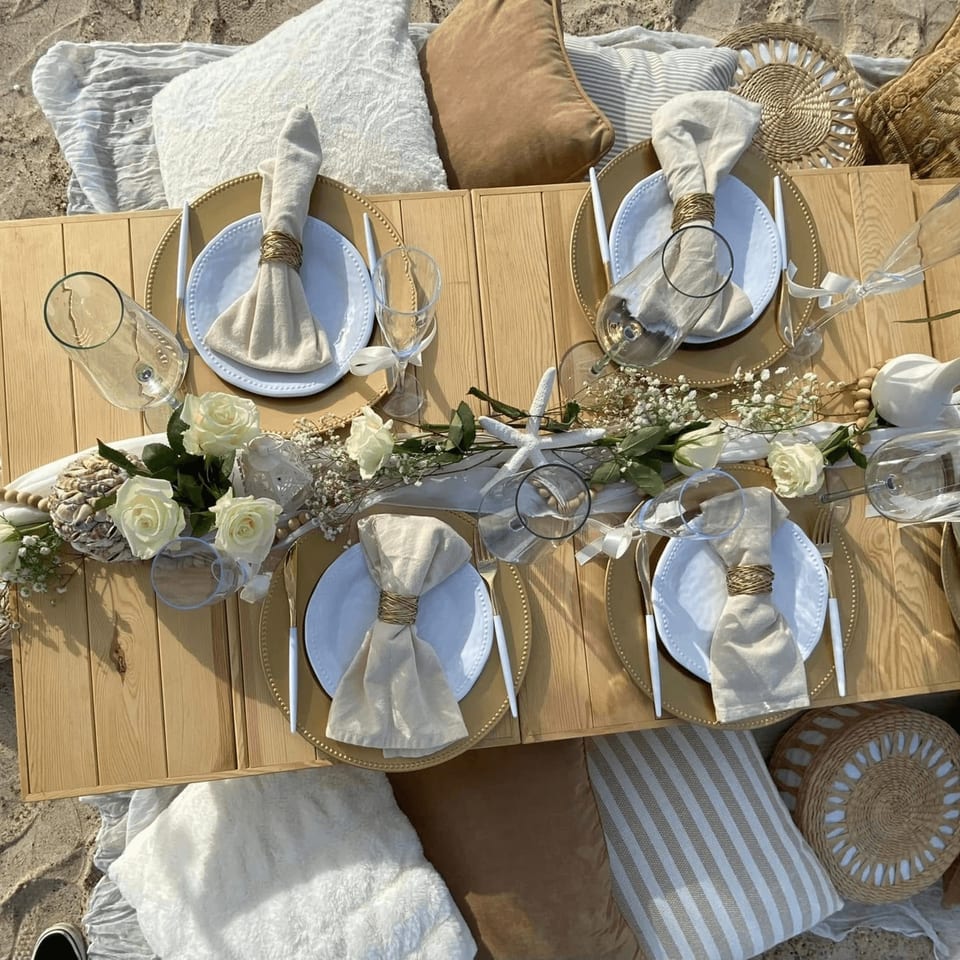 A beach picnic setup with a low wooden table, place settings, white roses, and pillows on the sand.