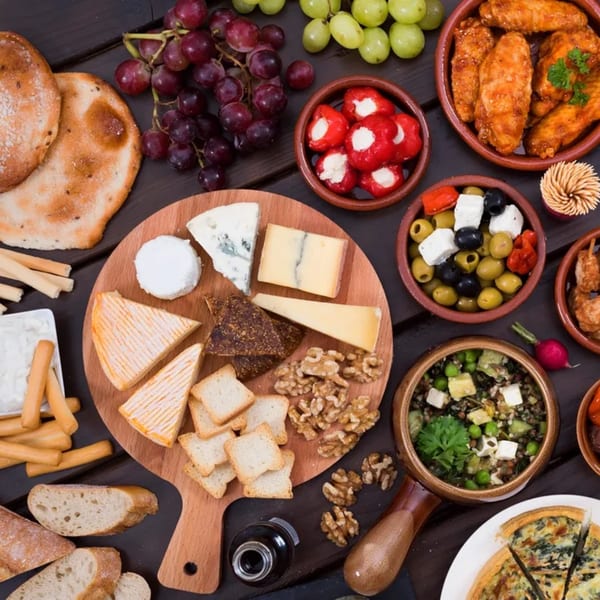 Overhead view of a deli spread with cheeses, bread, grapes, olives, peppers, and chicken wings.