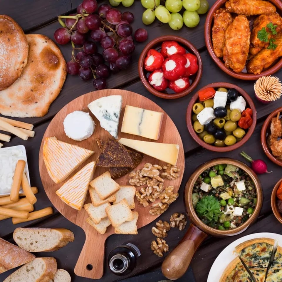 Overhead view of a deli spread with cheeses, bread, grapes, olives, peppers, and chicken wings.
