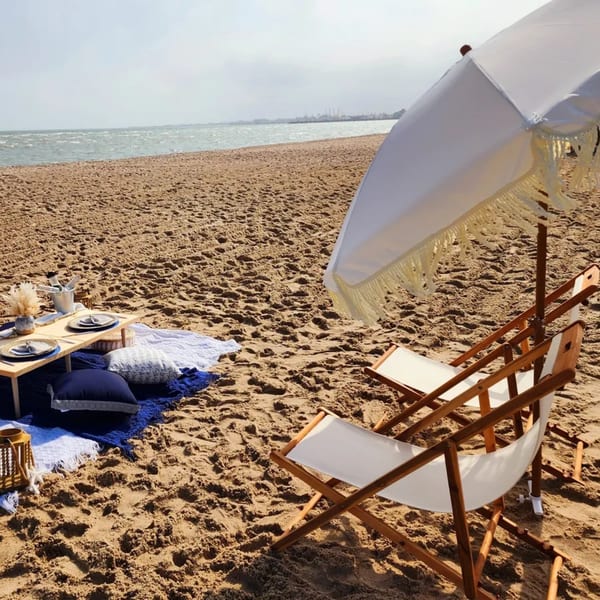 A beach picnic setup with a low table, cushions, and two lounge chairs under a white umbrella.