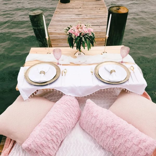 A romantic picnic setup on a wooden dock over water, with pink pillows and flowers.