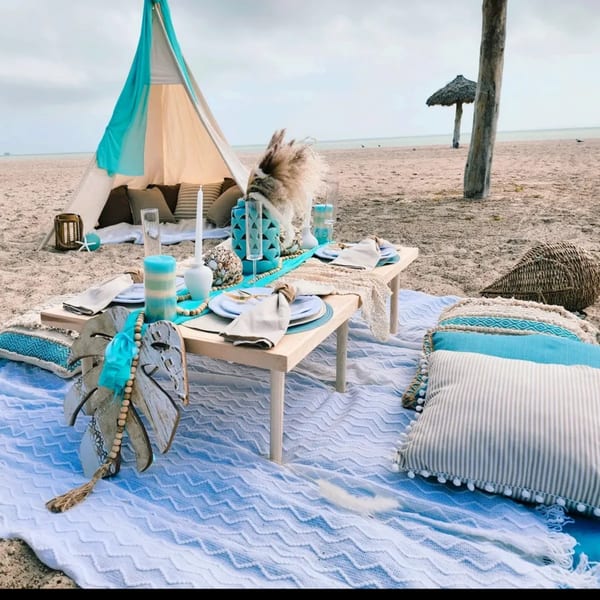 Boho-chic beach picnic setup with a teepee, low table, pillows, and turquoise decor on sand.