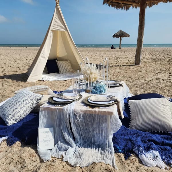 A beach picnic setup with a teepee, low table, pillows, and place settings on the sand.