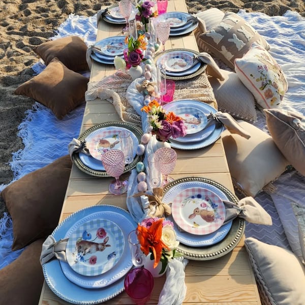 A picnic table set with bunny-themed plates, pink glasses, and flowers on a beach with pillows.