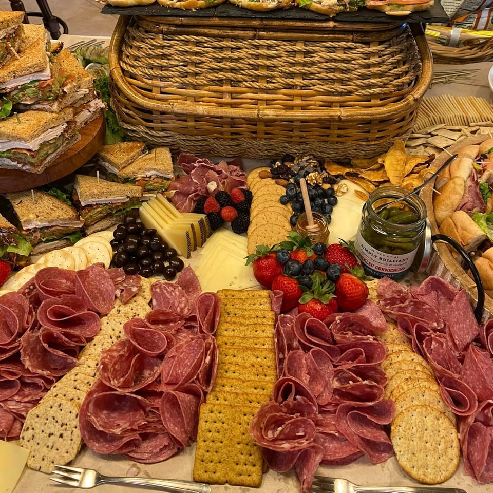 A large deli spread with sandwiches, charcuterie, cheeses, crackers, fruits, and pickled cornichons.