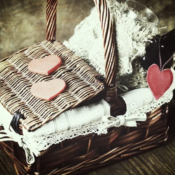 A wicker picnic basket with two heart-shaped decorations, a glass, and fabric inside.