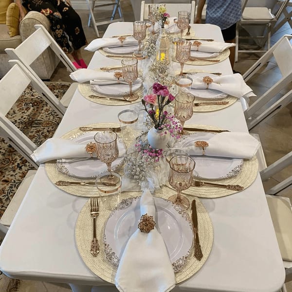 A long table set with white tablecloths, gold placemats, white plates, and pink floral centerpieces.