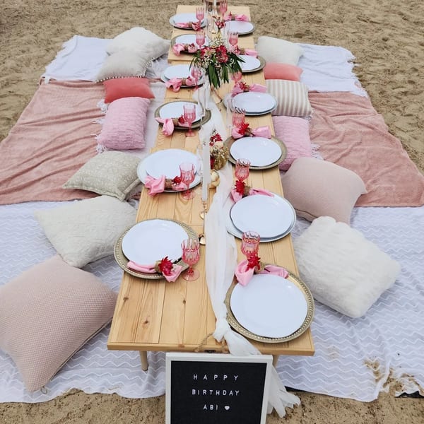 A picnic setup on sand with a low wooden table, white plates, pink napkins, and decorative pillows.