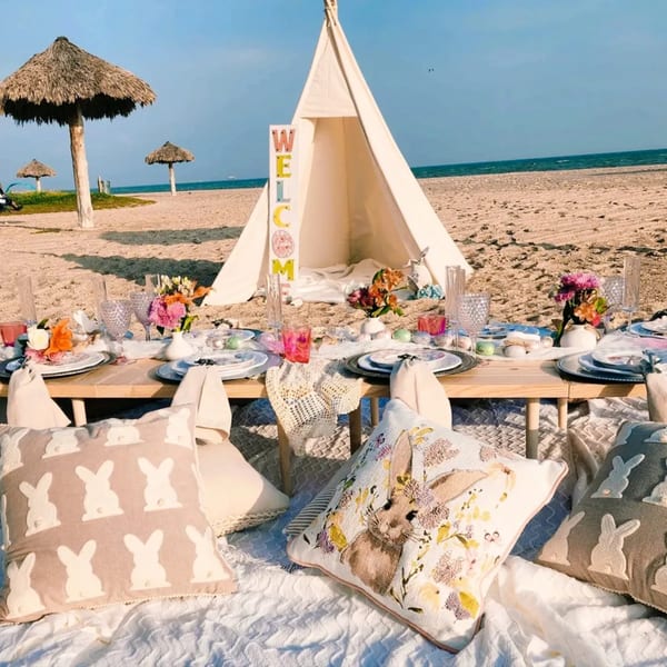 A beach picnic setup with a low wooden table, place settings, flowers, and bunny-themed pillows.