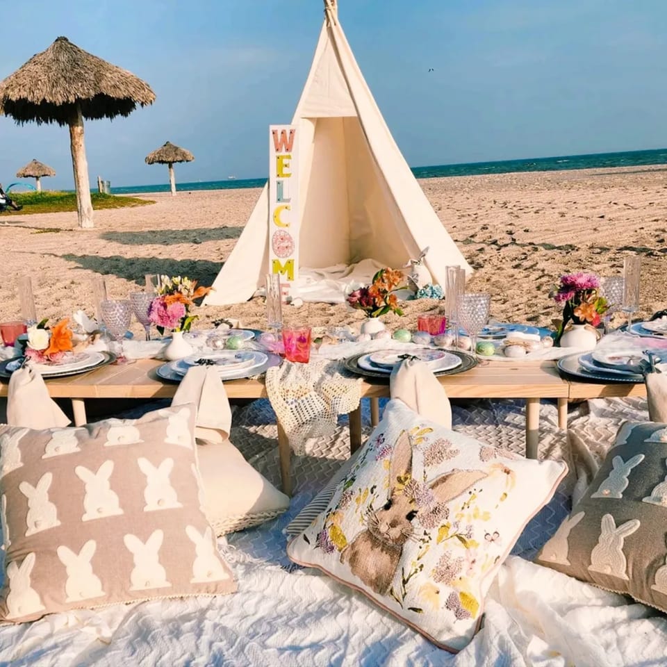 A beach picnic setup with a low wooden table, place settings, flowers, and bunny-themed pillows.