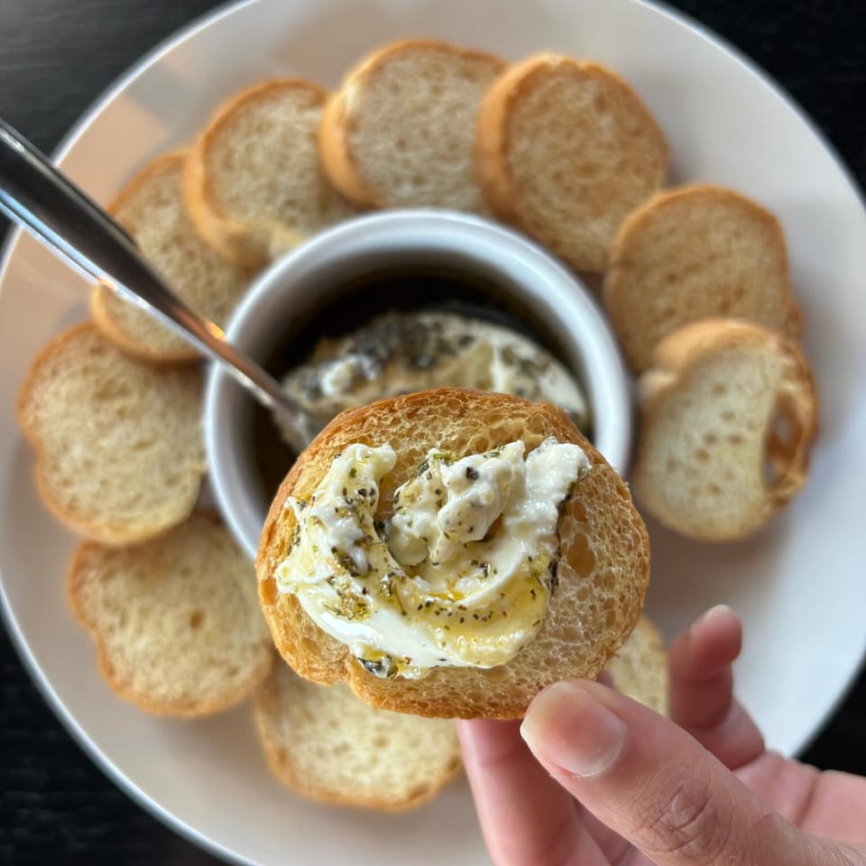 Hand holding a toasted baguette slice topped with creamy cheese, herbs, and honey, with a dip bowl.