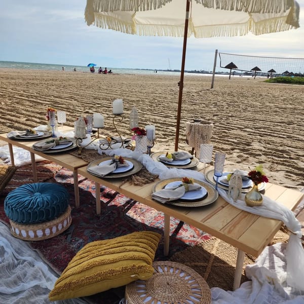 A beach picnic setup with a low wooden table, place settings, cushions, and an umbrella.