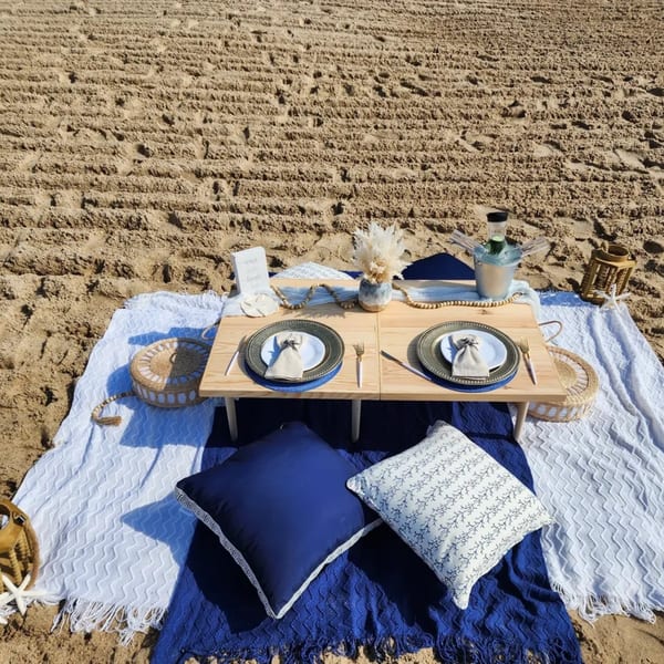 A beach picnic setup with a low wooden table, place settings, pillows, and blankets on the sand.
