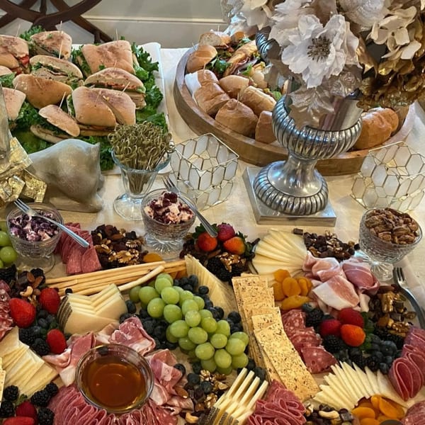 A deli spread with sandwiches, charcuterie, cheeses, fruits, nuts, and crackers on a white tablecloth.