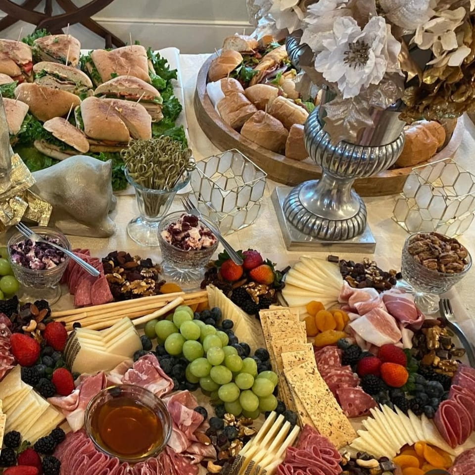 A deli spread with sandwiches, charcuterie, cheeses, fruits, nuts, and crackers on a white tablecloth.