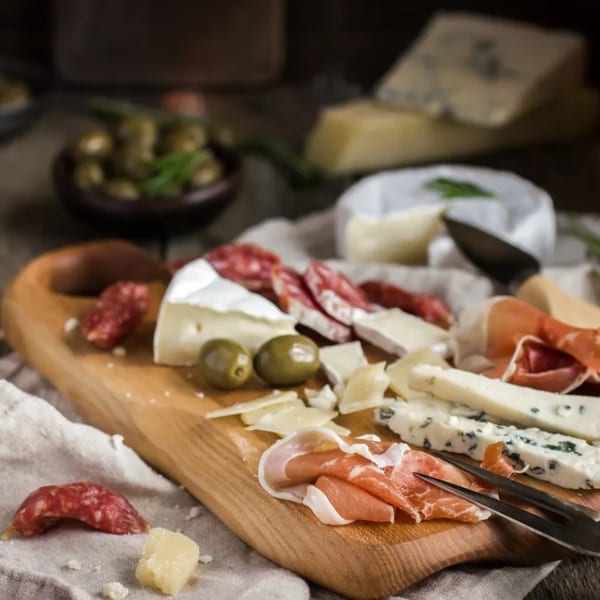 A wooden board with various cheeses, sliced salami, prosciutto, and green olives.