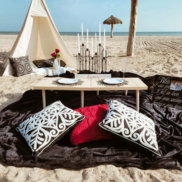 Romantic beach picnic setup with a teepee, low table, pillows, candles, and ocean view.