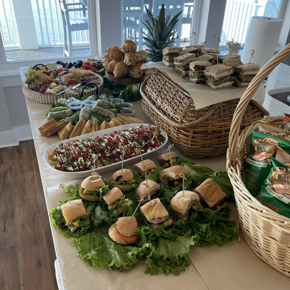 A deli spread with sandwiches, charcuterie, salad, and chips on a table by a window.