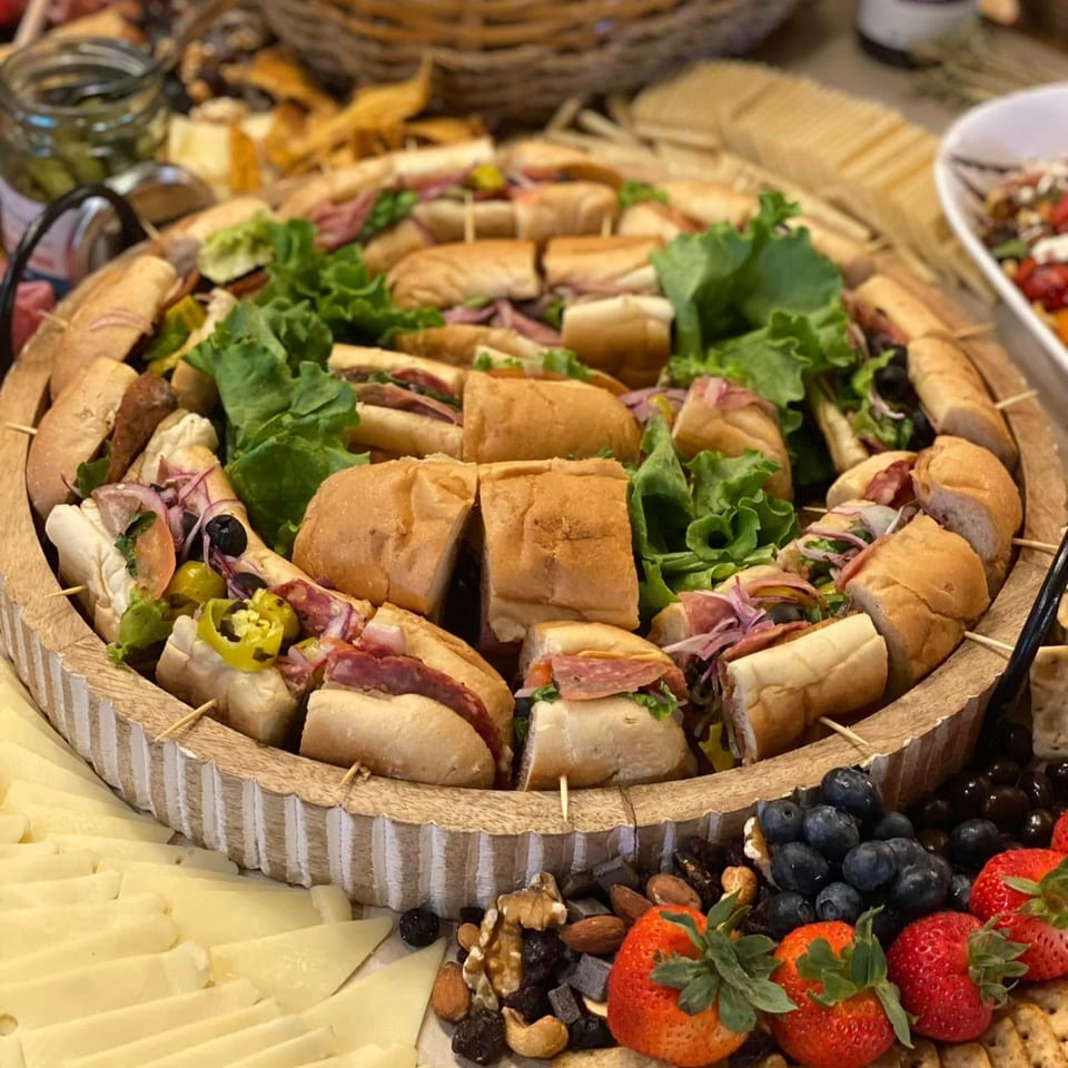 A large wooden platter filled with various deli sandwiches, cheeses, fruits, and nuts.